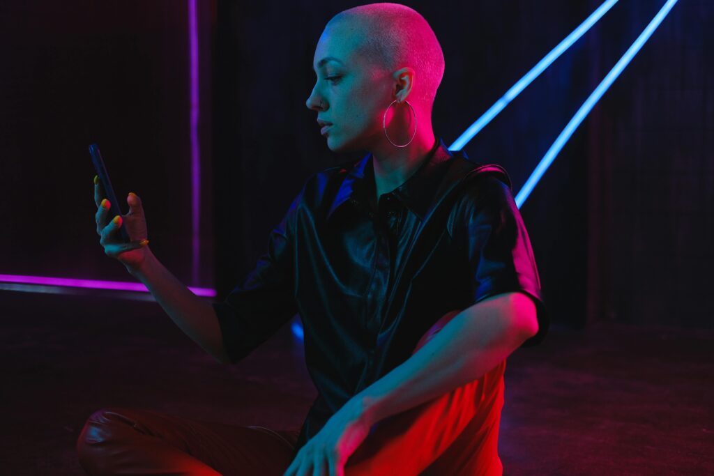 Woman with shaved hair sitting on floor in night club, looking at phone under neon lights.