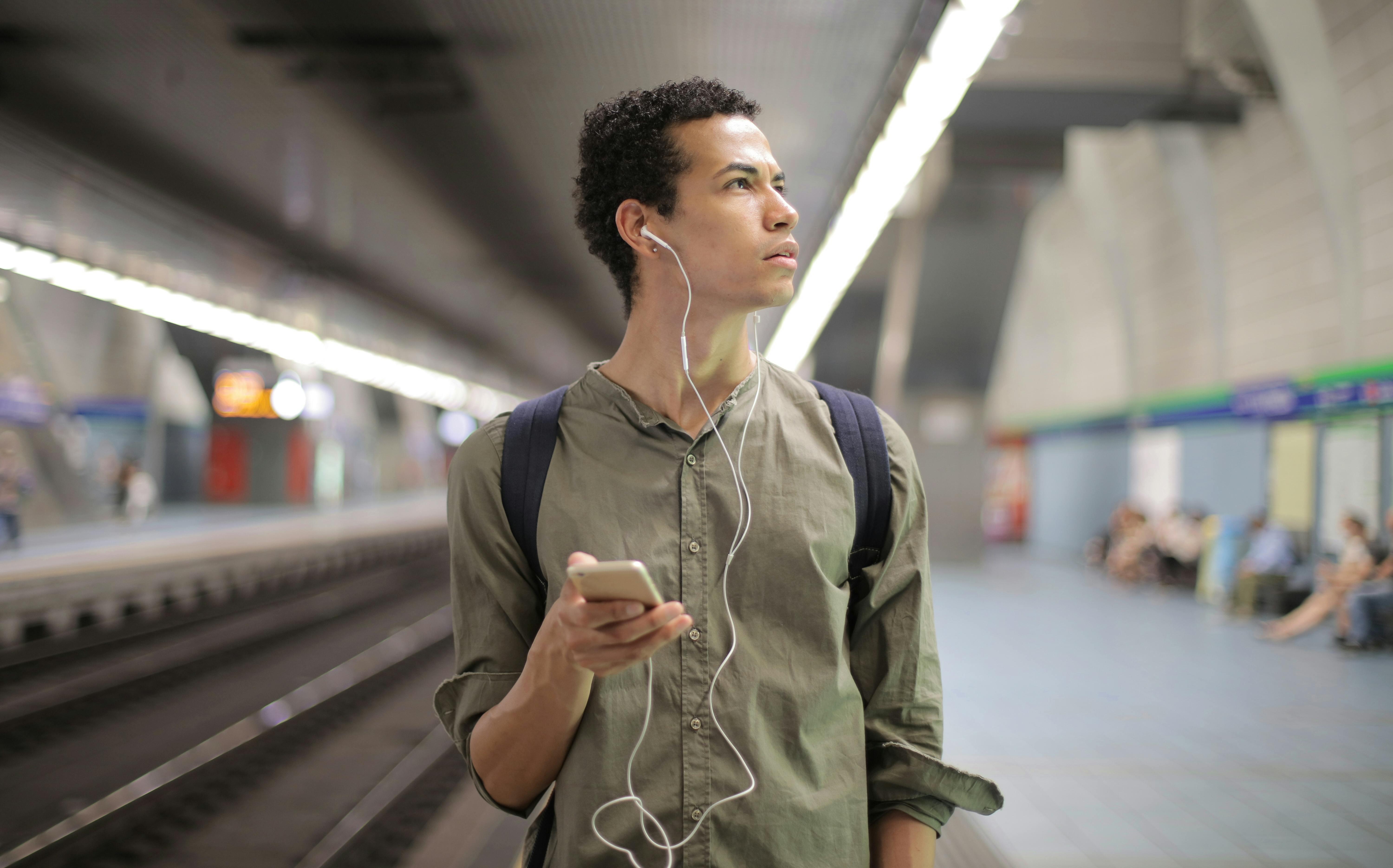 A man standing in a train station, wearing earphones.