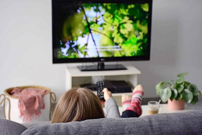 Woman sitting on a couch pointing a remote control at a television displaying captions
