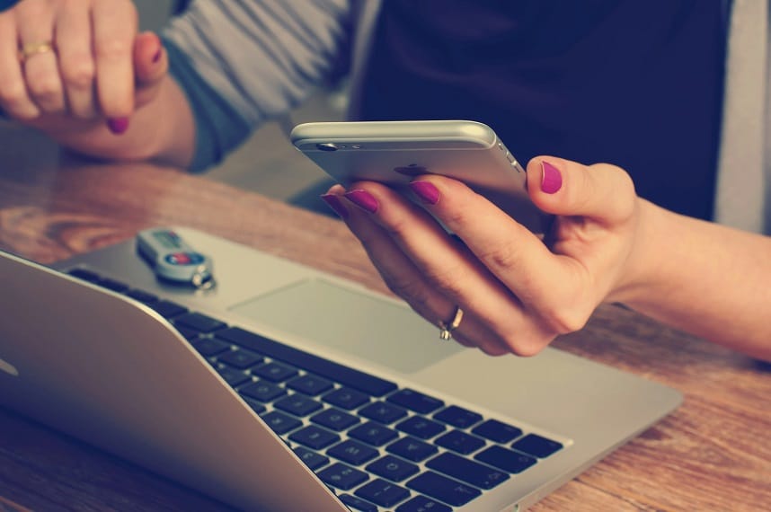 A woman holds a cell phone in her hand. She is working at a table above an open laptop