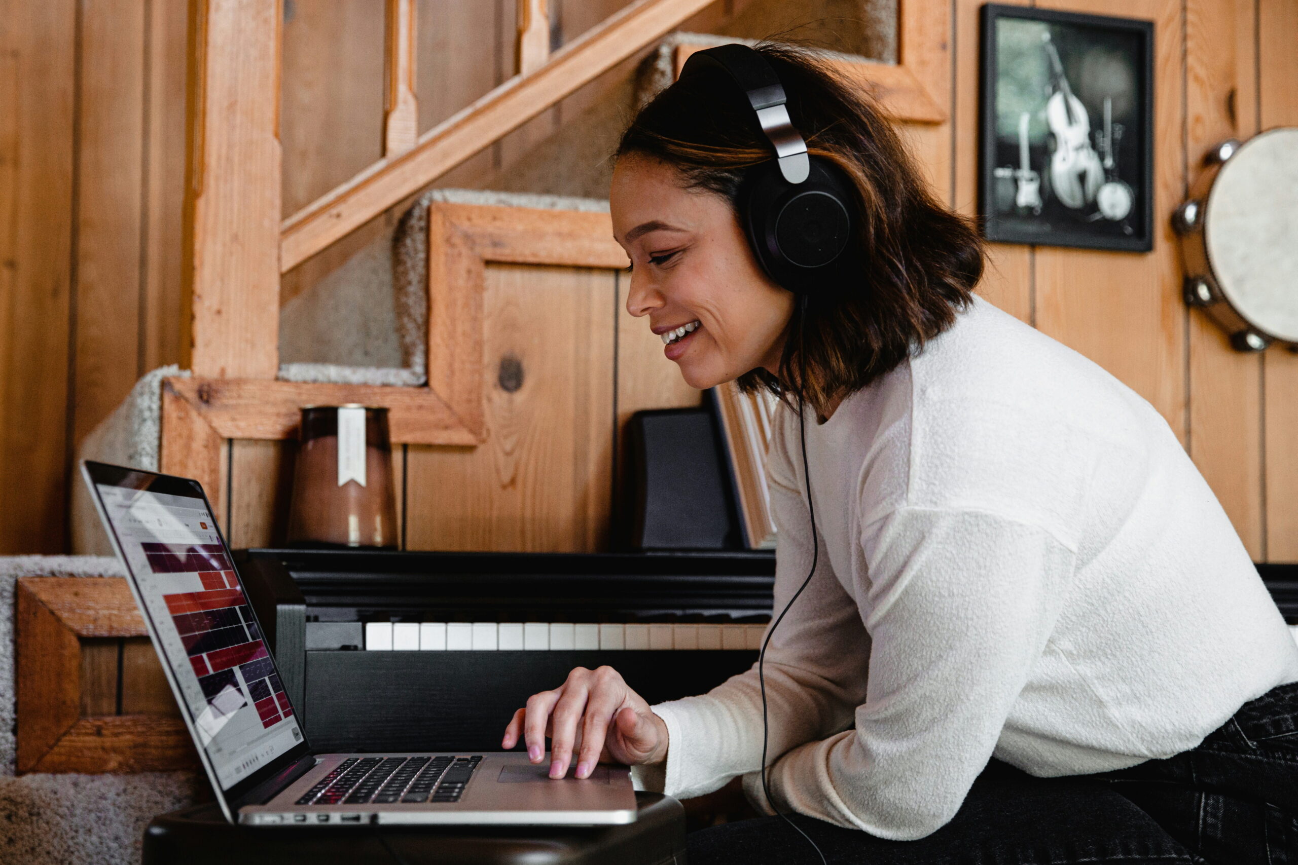A smiling woman wearing headphones typing on a laptop computer with a piano and wooden stairs in the background