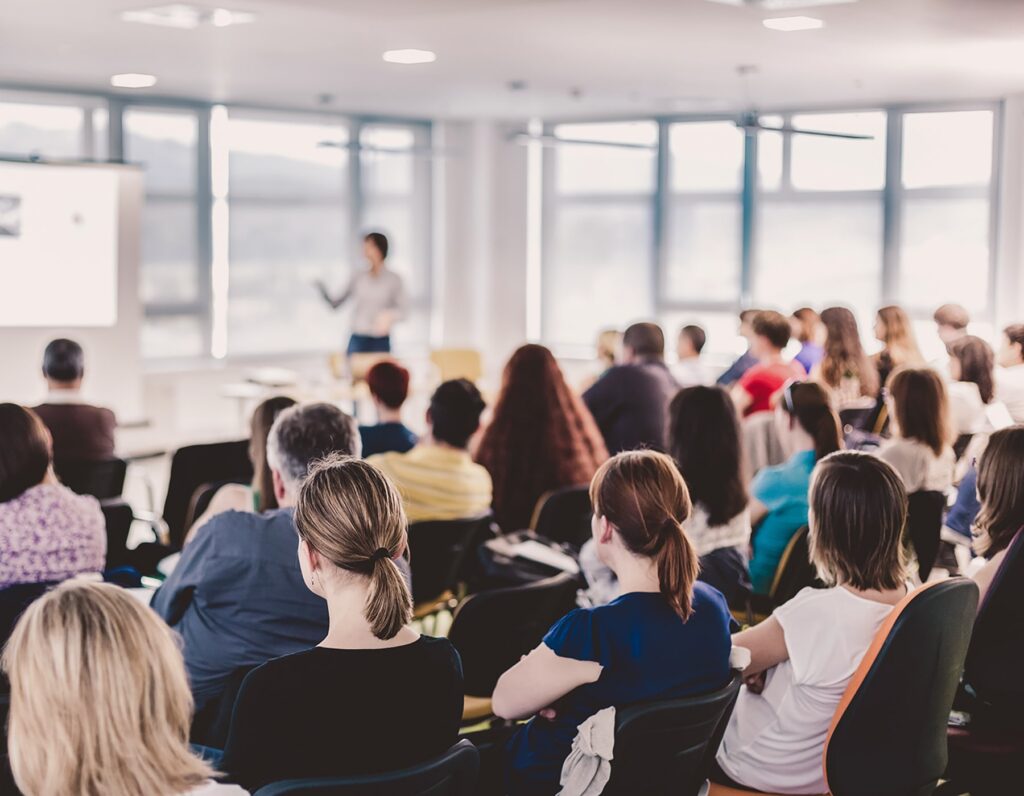 A conference room is shown full of seated attendees who listen as a speaker talks