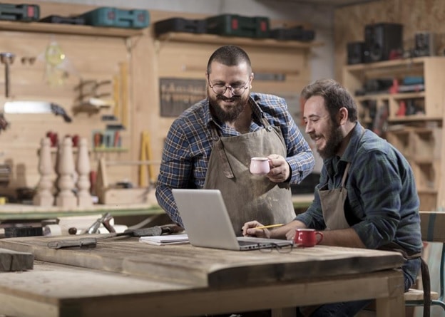 Two men in a wood-working shop, wearing work aprons and looking at an open laptop