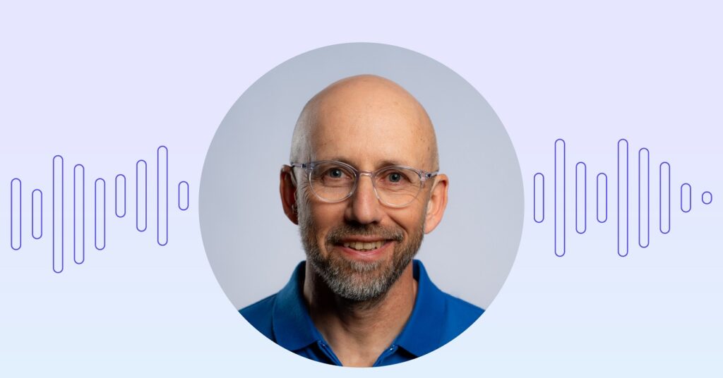 Yair Amsterdam, a man wearing a blue shirt and glasses is pictured in front of a white background