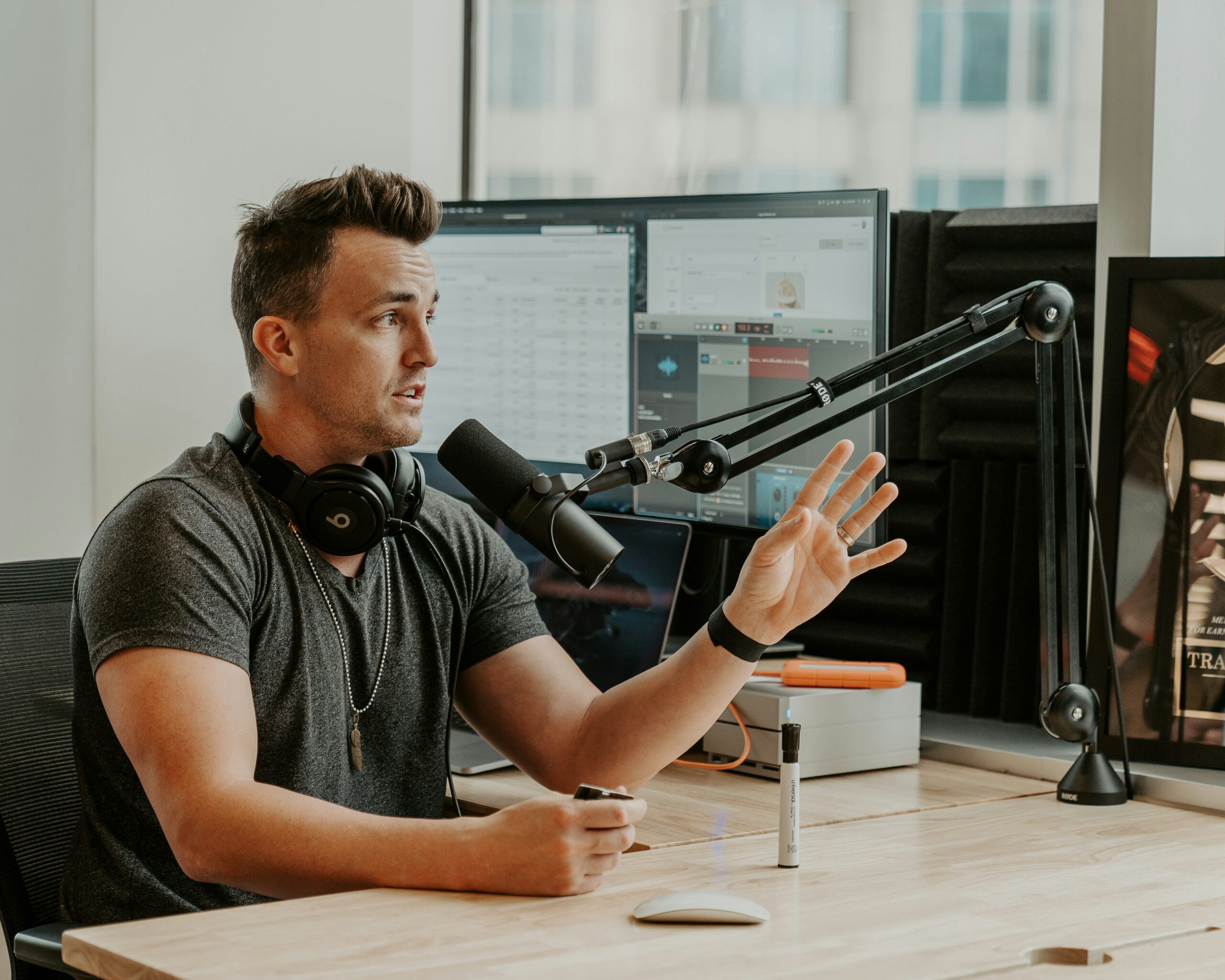 Man wearing grey T-shirt sitting by a desk using a microphone while podcasting with headphones around his neck