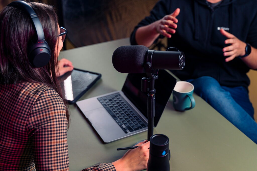 A female podcast host and guest having a discussion with headphones and microphone and a laptop visible.