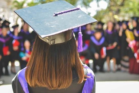 Photographed from behind and over the shoulder, a woman wearing a graduation cap and gown walks towards other graduates.