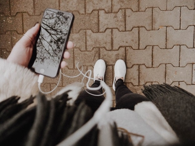 Shot from the point of view of a woman looking down at her cell phone, the woman holds a phone in her left hand and we can see white headphone wires and a white pair of shoes 