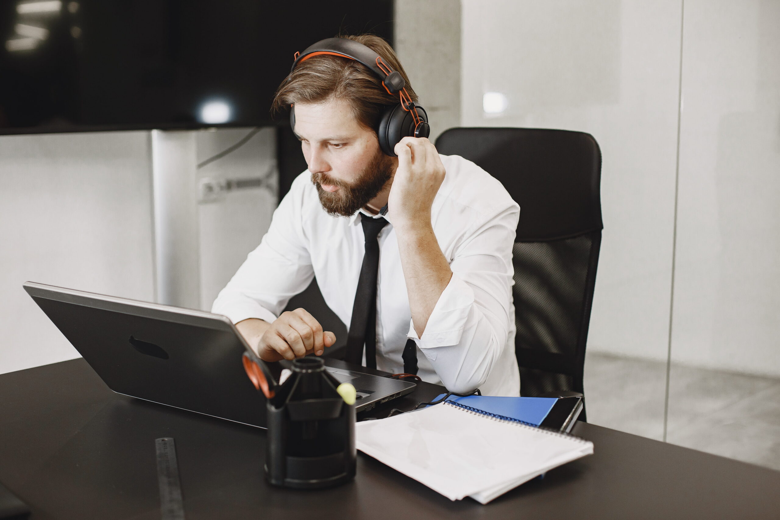 A bearded businessman wearing a white shirt, black tie, and headphones sitting at a desk and looking intently at an open laptop.