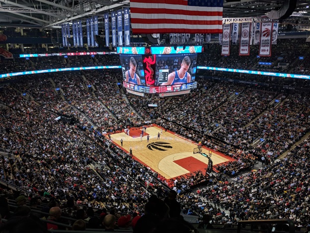 An overhead shot of a basketball game inside a filled arena. The scoreboard can be seen hanging above center court
