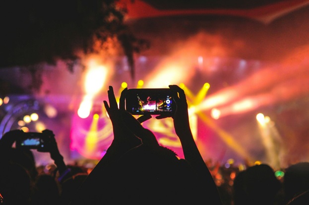 Silhouetted against a lighted stage, a person holds up a cell phone at a concert and records