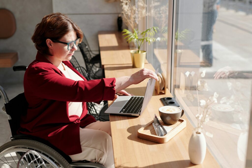 A woman in a red sweater sits in a wheelchair at a desk and uses a laptop computer