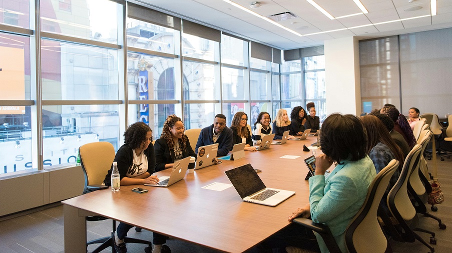 Employees sit at a long table in a corporate boardroom, a wide, tall window is in the background