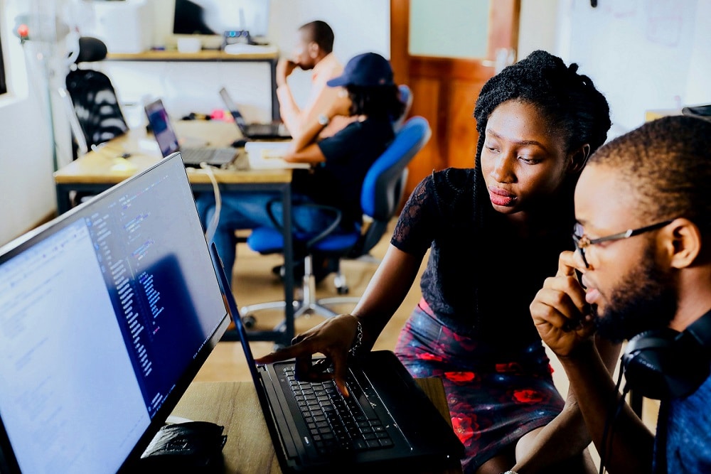 Two students sit at a computer. One student - a woman - is pointing to the screen while the other - a man - looks on.