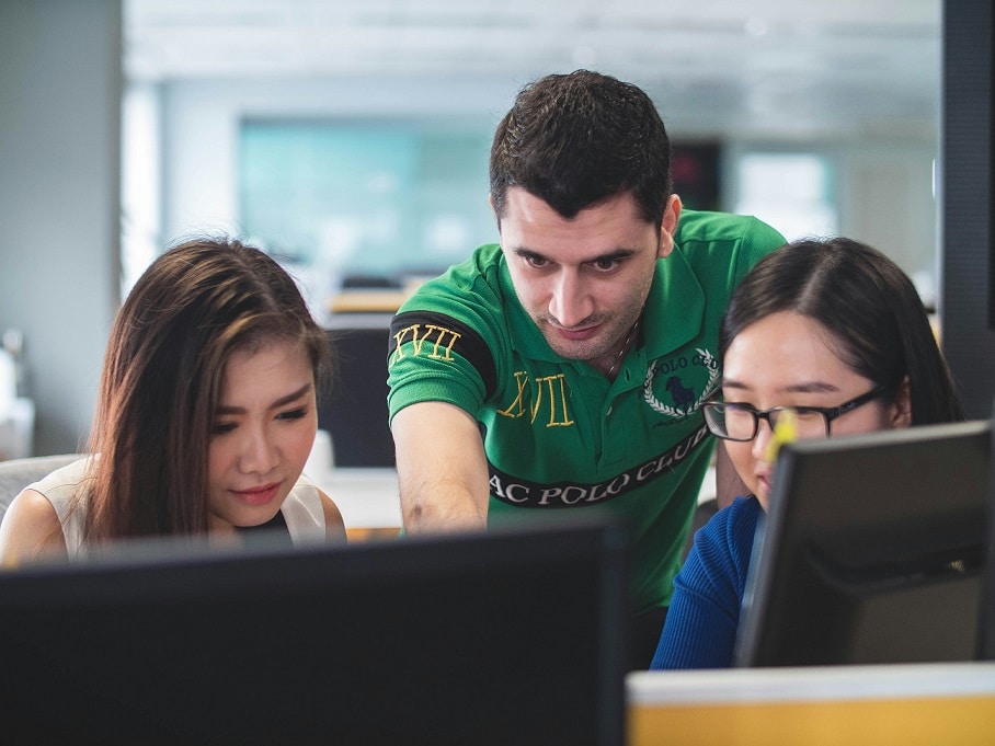 Three students - two women and a man - works at a computer station. We see them from behind the computers looking towards the camera.