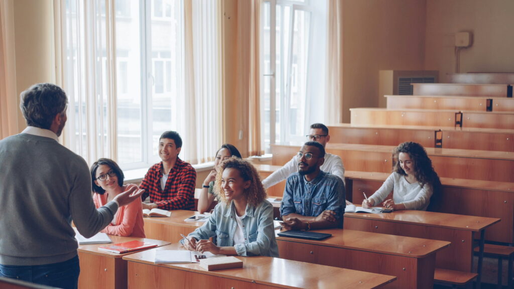 A mature male professor facing a diverse group of attentive university students seated at wooden desks in a bright lecture hall, with large windows in the background.
