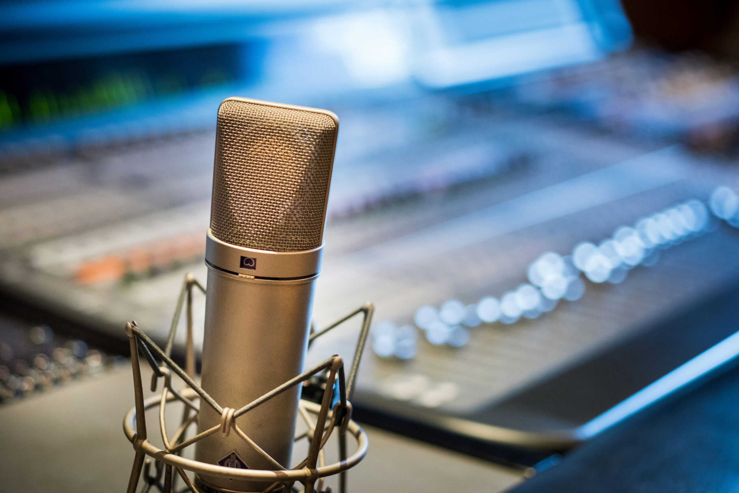 A close-up of a professional condenser studio microphone in a shock mount, with an audio mixing board blurred in the background.