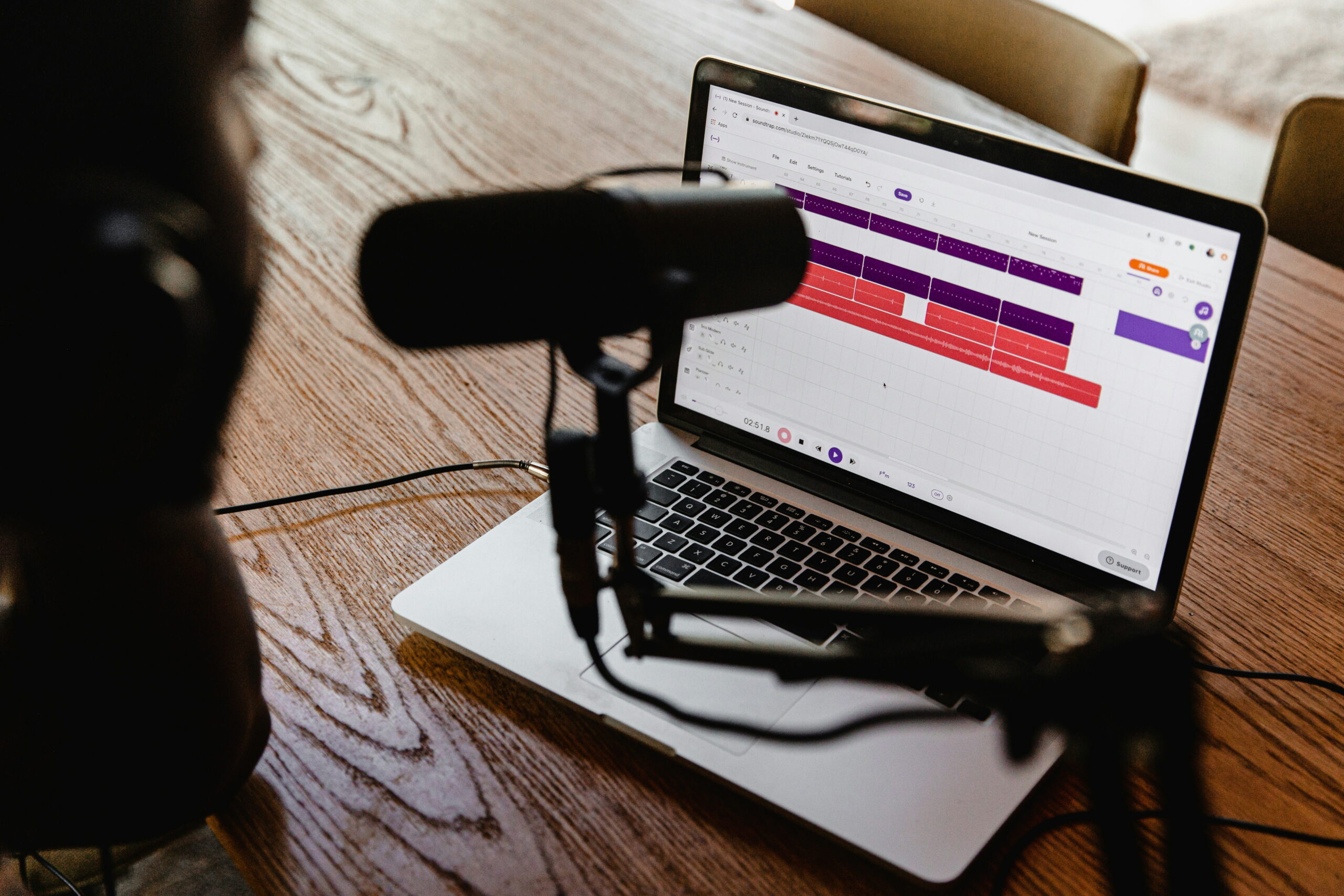 A person wearing headphones sitting at a wooden table with a microphone on a boom arm facing a laptop displaying an audio waveform editing software.