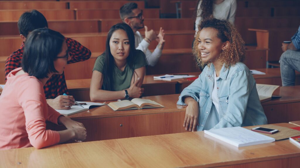Three female students and one male student are seen talking in a circle with books open in a lecture hall with more students visible in the background. 