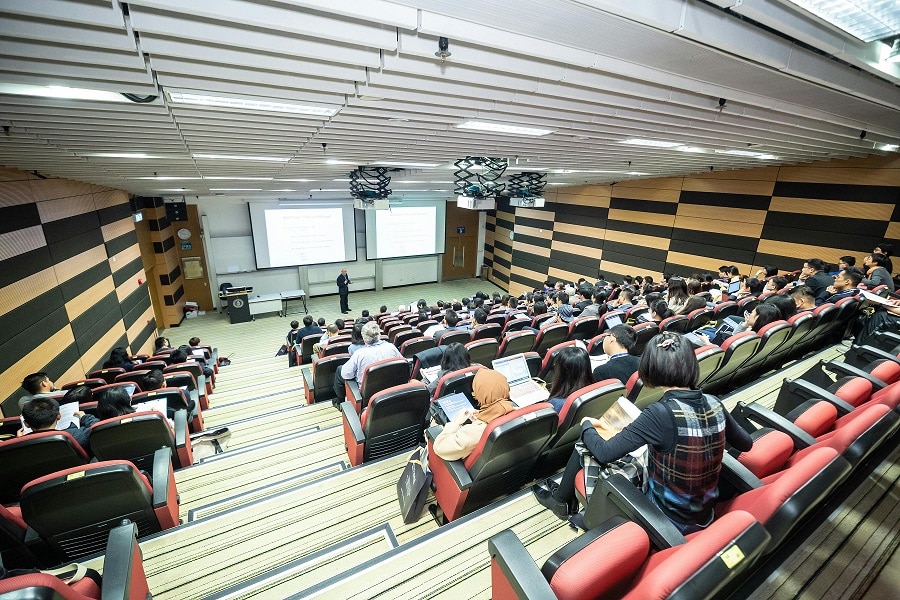 Students sit in a lecture hall, a person stands in front in front of two large white screens