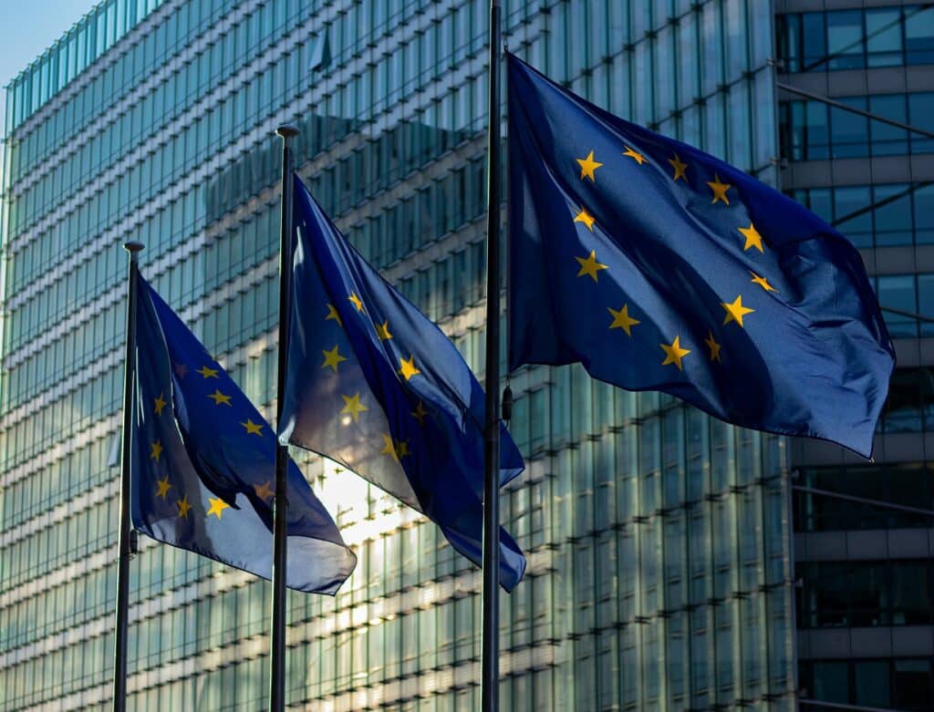 Three European Union flags fly in front of a plate glass building facade. The flags are blue with a circle of yellow stars.