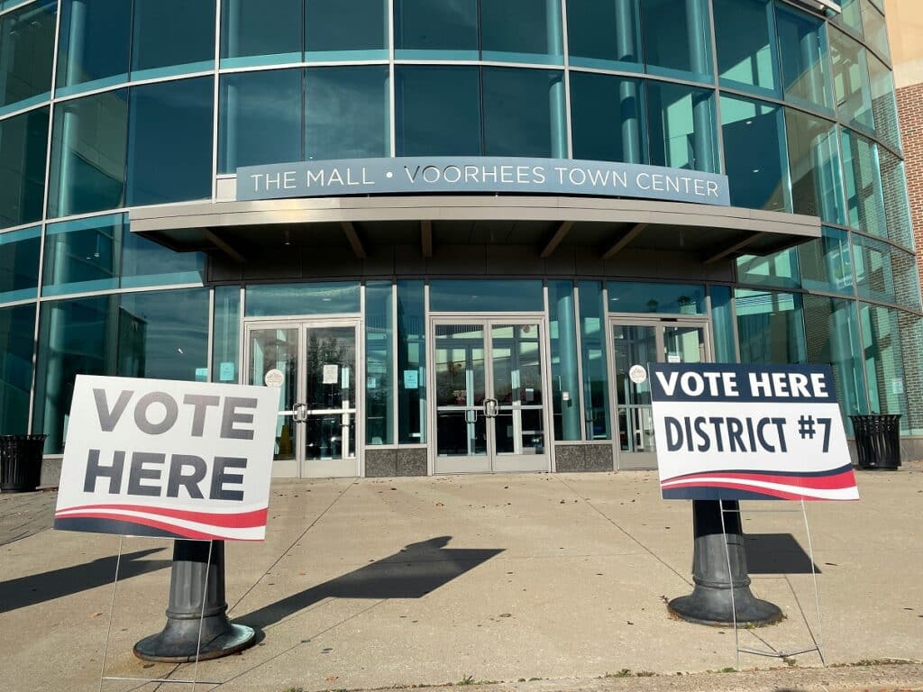 Two election signs - one reading 'vote here' and another reading 'vote here - District 7' sits in front of large glass doors outside of the Voorhees Town Mall