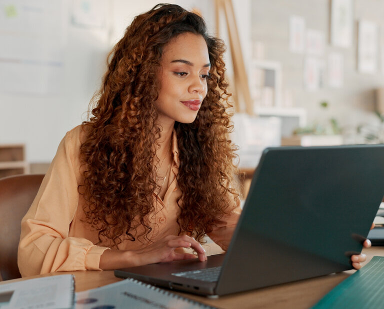 A woman with dark, curly hair wearing a peach-colored shirt sits at a desk and scrolls on an open laptop.