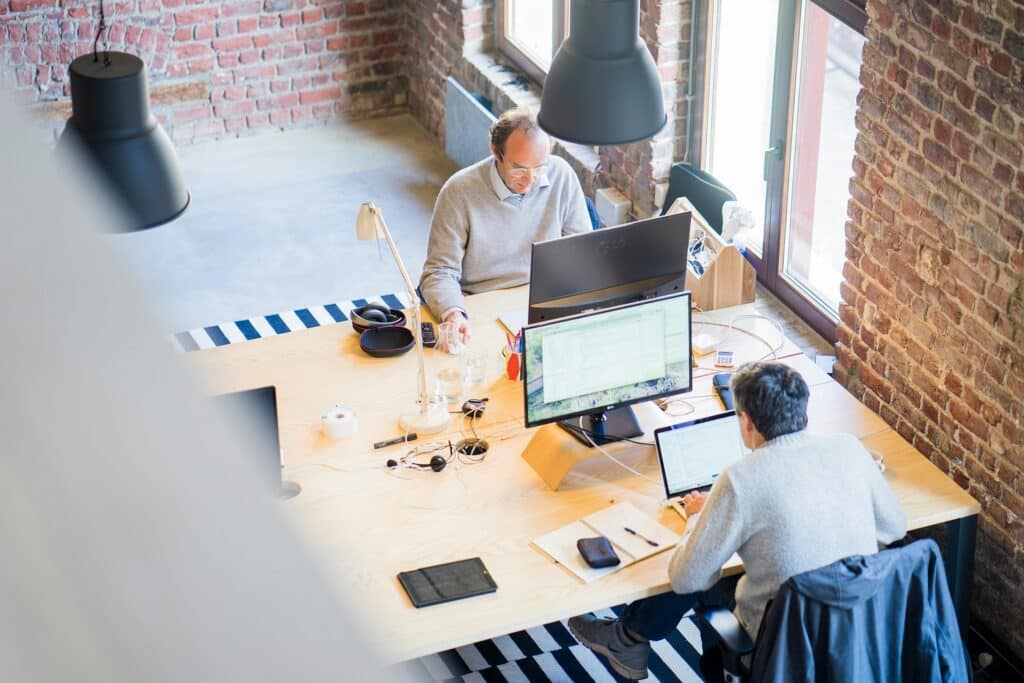 Two men work at two computer stations at a shared desk in an office.