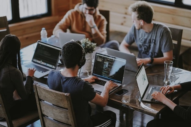 Three young men and one young woman work on laptops at a large square table.