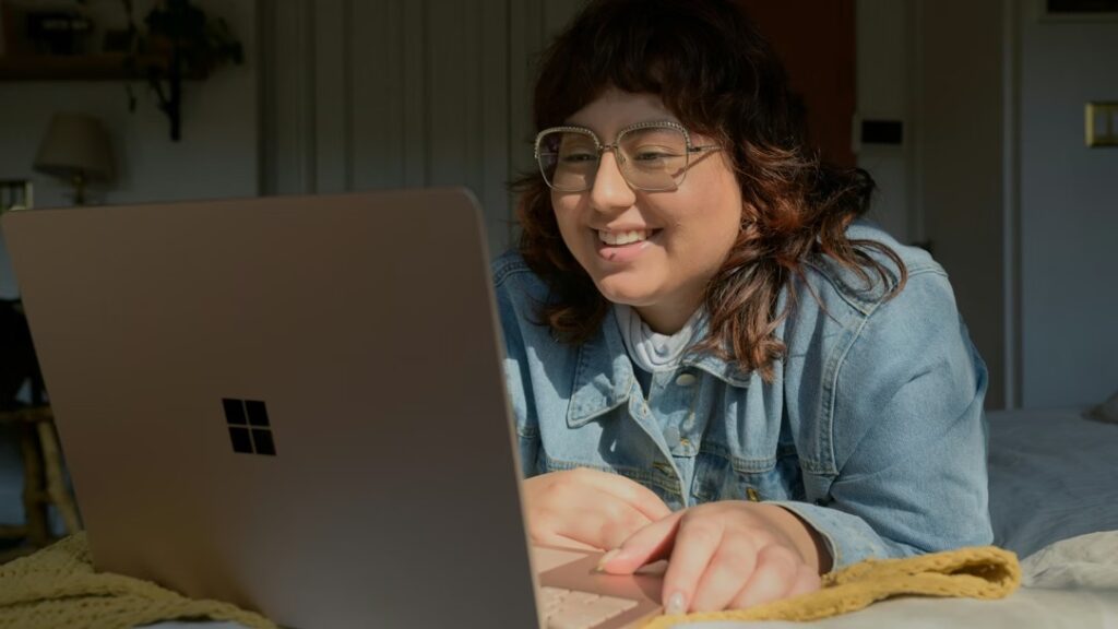 A woman with dark hair and wearing a light blue shirt looks at the screen of her open laptop.