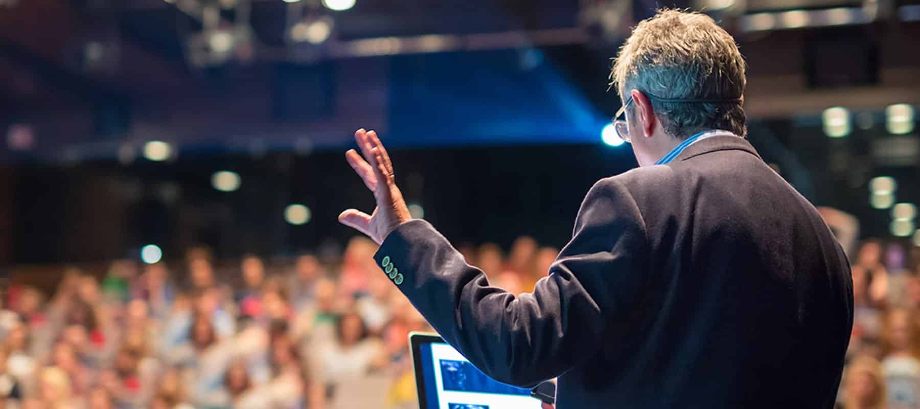 A middle aged man gives a lecture on stage in front of an audience while wearing a suit.