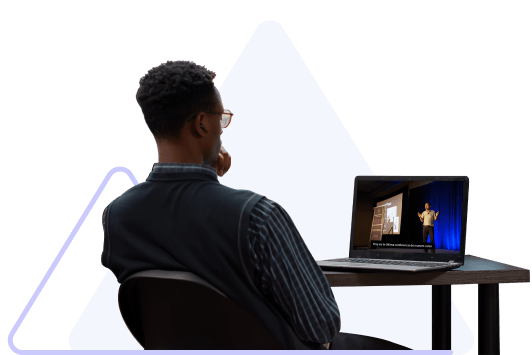 A man with dark hair and glasses wearing a dark-colored suit sits with his back to the viewer and watches a video on his laptop.