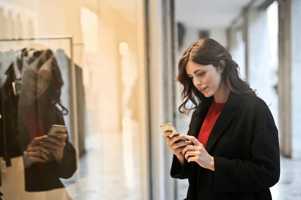 A woman with brown hair wearing a red shirt and black jacket stands outside a store and looks at her cell phone.