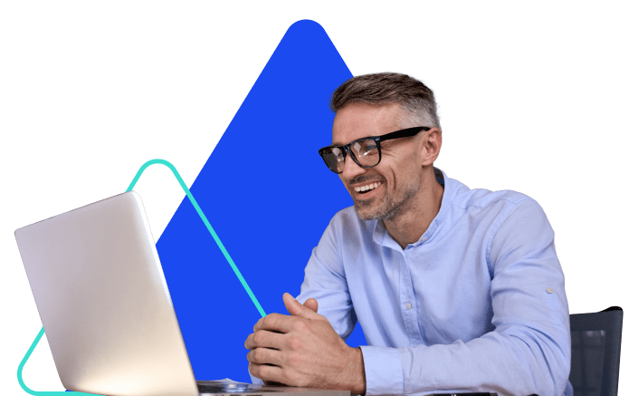 A man wearing glasses sits at a desk and looks at his laptop screen.