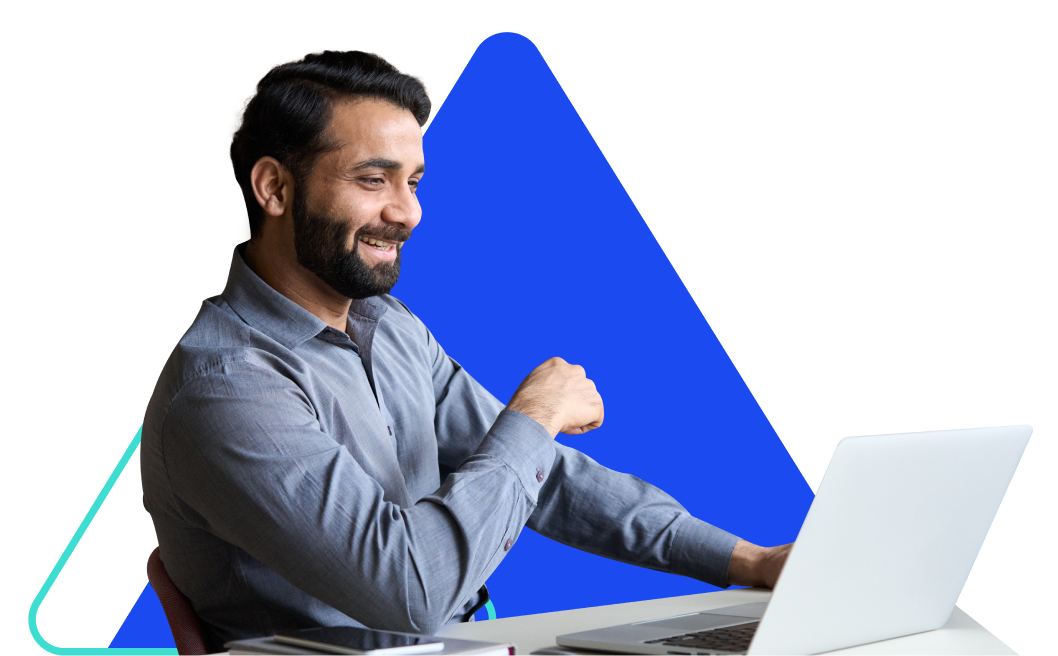 A smiling man with dark hair and a blue shirt sits at a desk and looks at his laptop.