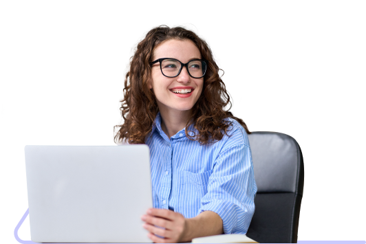 A woman with reddish-brown hair wearing glasses and a light blue shirt looks to her left as she works on a laptop.