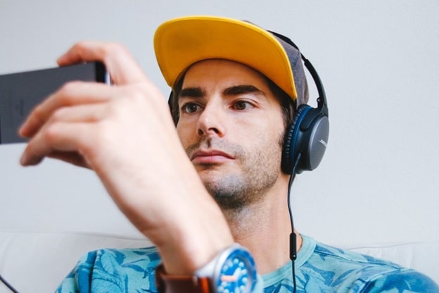 A man wearing a yellow-billed baseball cap and headphones watches a video on his cell phone.