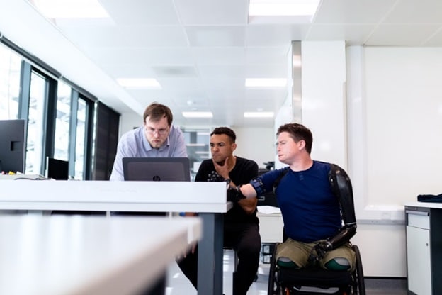 Three men - one with a prosthetic arm and prosthetic legs, sitting in the wheelchair - look at a laptop in an office.