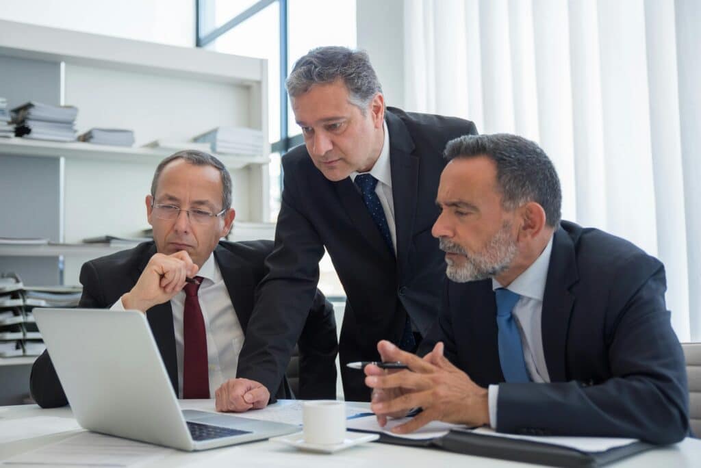 Three men in suits and ties gather around a desk and look at the screen of an open laptop.