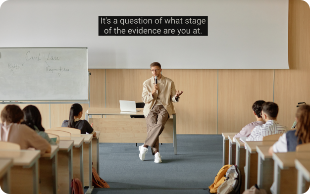 A man with light brown hair and glasses wearing tan pants and a tan jacket holds a microphone and sits against a table in front of a classroom. Captions at the top of the image read: 