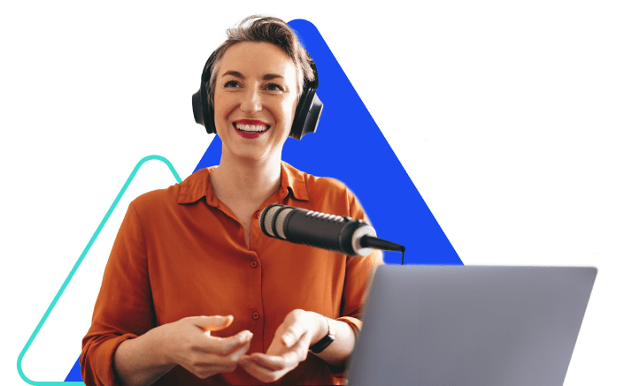 A woman wearing headphones and an orange blouse stands in front of a microphone and open laptop.