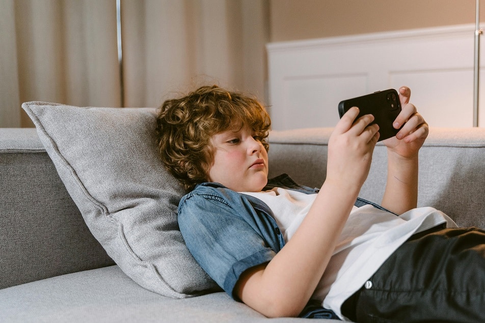 A boy with curly hair rests against a pillow on a couch and looks at his phone