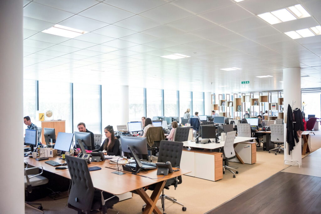A wide view of a bright, modern open-plan office space filled with wooden desks, computer monitors, and employees working during the day.