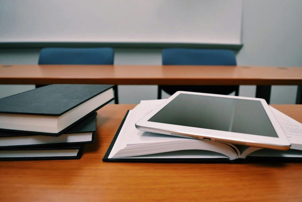 Close up of a desk in a school classroom. A tablet sits atop an open text book on the desk. 