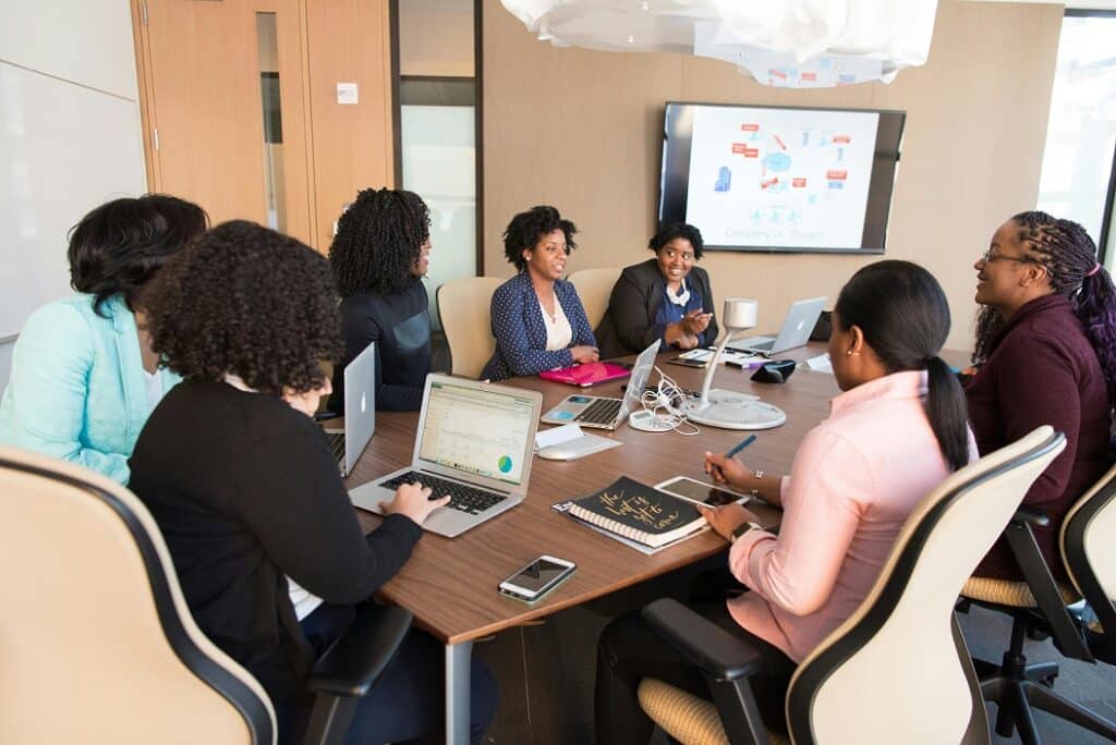 Professionals are seated around a table in a conference room. Laptops and tablets can be seen on the table.