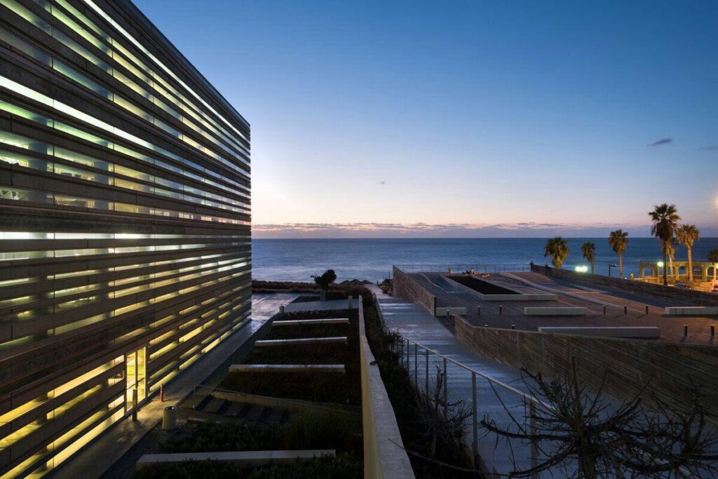 Modern building with horizontal glass panels illuminated at dusk, overlooking a landscaped terrace and the Mediterranean Sea with palm trees along the waterfront.
