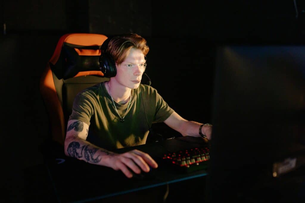 A young man with reddish hair and wearing an olive green shirt sits in front of a desktop gaming set-up.