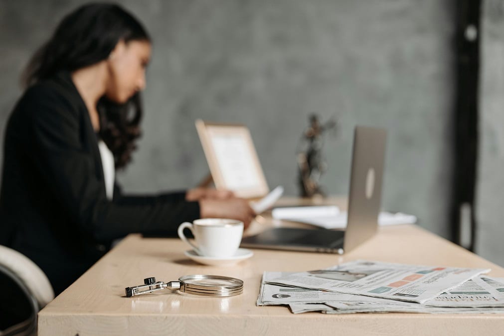 A woman wearing a black blazer sits at the end of a long table and works on an open laptop.