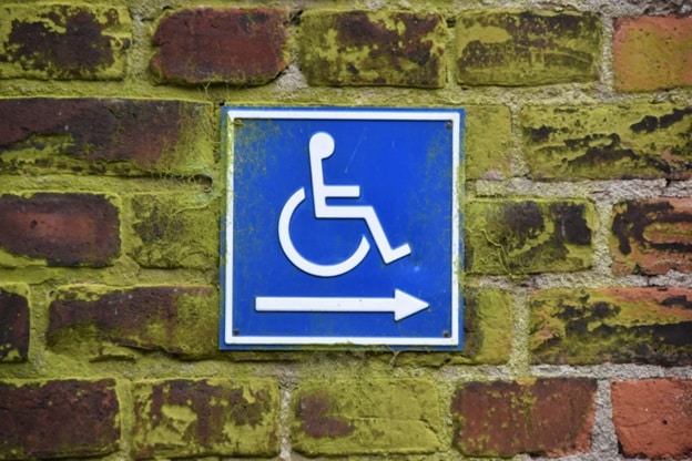 A blue and white handicapped accessibility sign is posted against a greenish-colored brick wall.