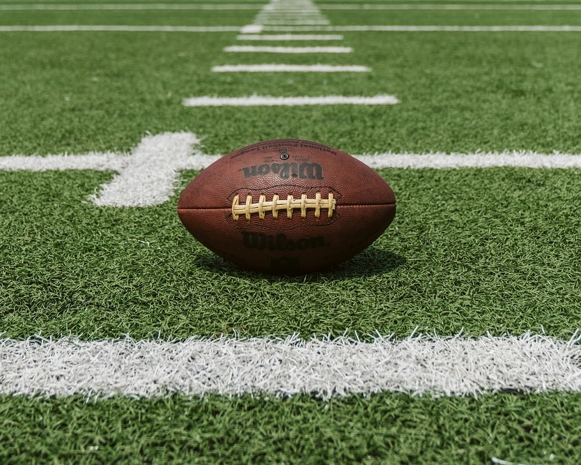 A brown leather football with the company name 'Wilson' sits on the white hashmarks of a green artificial turf football field